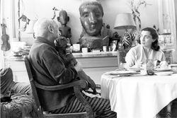 Jacqueline and Pablo Picasso seated at the dining table; a dachshund, in Picasso's lap, tries to lick the artist's face.1957